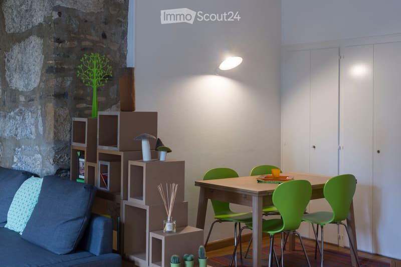 A modern, cozy living space with a stone accent wall, a grey sofa with patterned cushions, a wooden dining table with green chairs, and a modular shelving unit with decorative items.