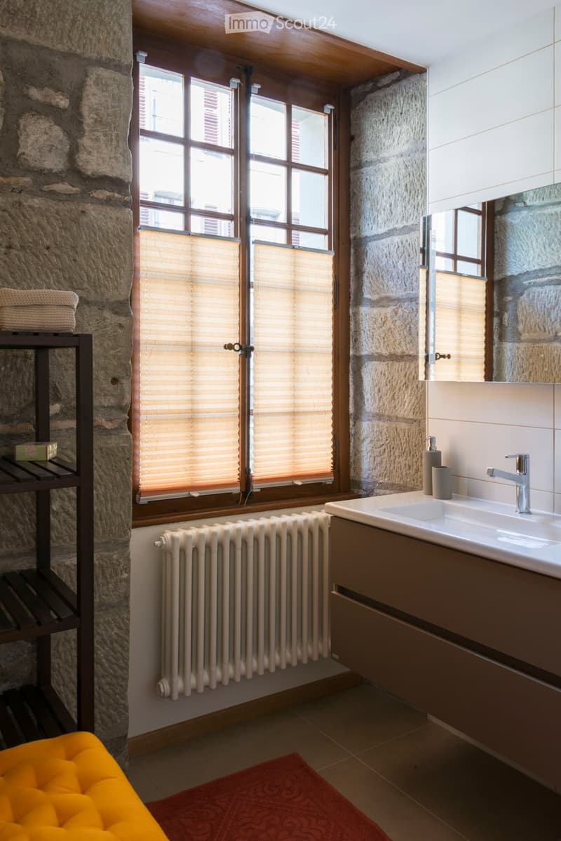 Bathroom with stone walls, a window with blinds, a sink with a modern faucet, a radiator, a mirror, a small cabinet, and a yellow stool with a red rug.