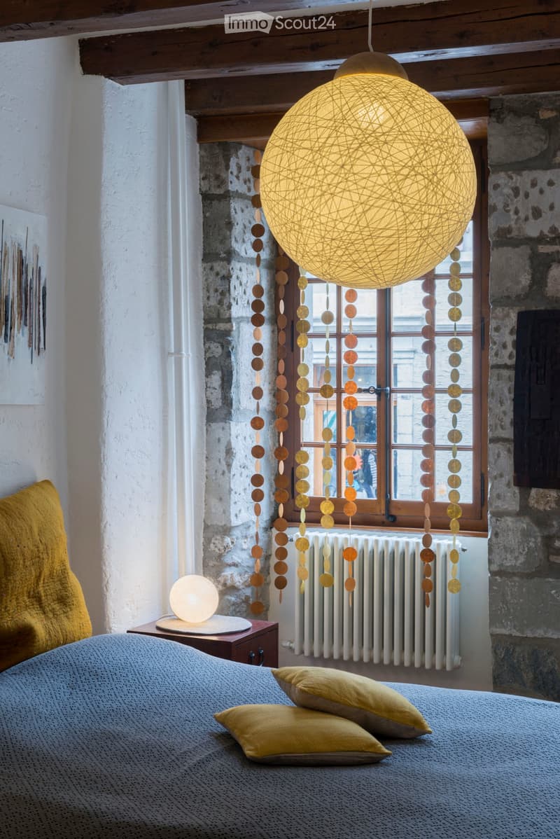 Cozy bedroom with a grey duvet, yellow pillows, a wooden nightstand with a table lamp, exposed wooden beams, stone walls, and a large window with hanging circular decorations.