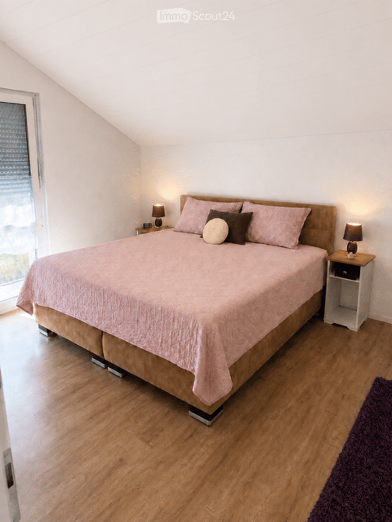 Bright bedroom with wooden flooring, a large bed with pink bedding, two nightstands with lamps, and a purple rug in the corner.