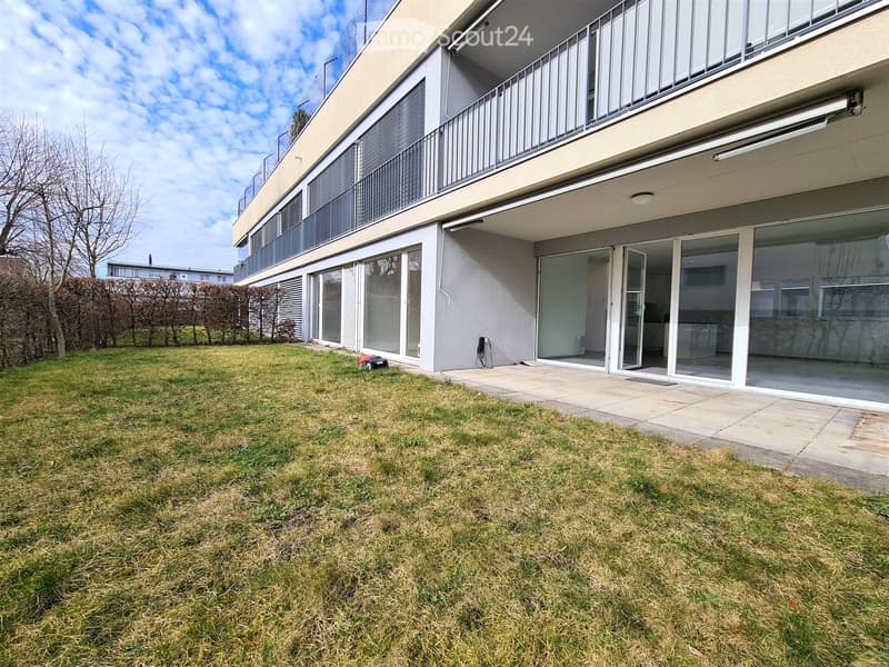 Lawn area in front of a modern building with large glass windows and a balcony, surrounded by hedges and trees.