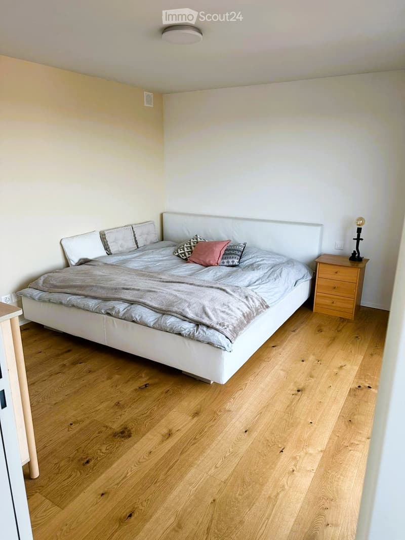 Modern bedroom with a white bed, grey bedding, wooden nightstand, and a lamp. Light wooden flooring and white walls.