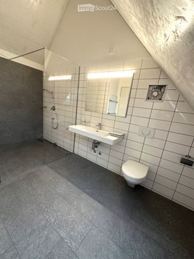 Modern bathroom with white tiled walls, grey floor, glass shower enclosure, sink, mirror, and toilet.