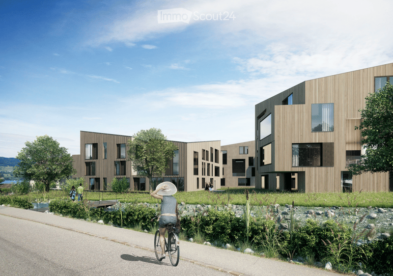Modern multi-story residential building with large windows and wooden facade, surrounded by greenery and a pathway.
