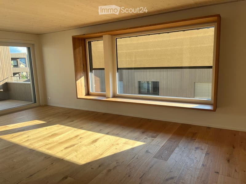 Empty room with wooden flooring, large windows with blinds, and a sliding glass door leading to a balcony.