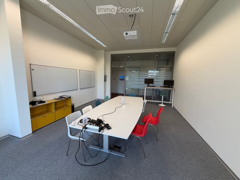 Modern conference room with a long white table, several chairs, a projector screen, and a small cabinet with shelves.
