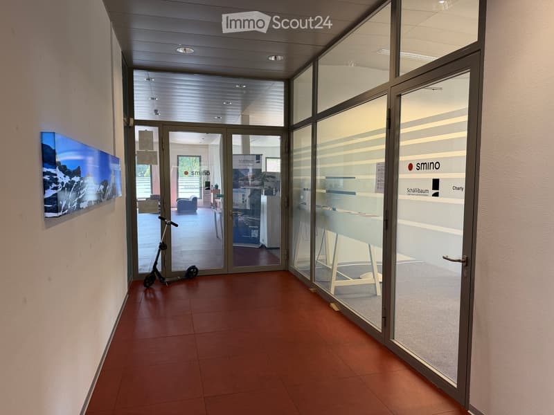 Corridor with glass doors leading to an office area, featuring a scooter parked near the entrance and a large wall-mounted display on the left side.
