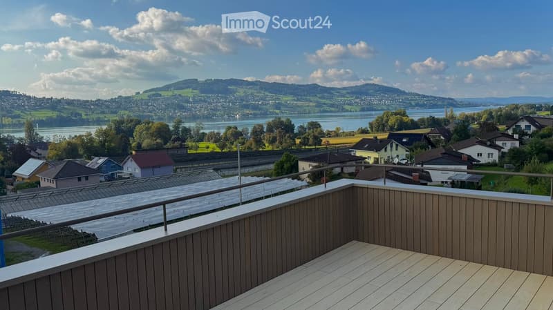 Balcony with wooden decking and metal railing, overlooking a scenic lake and town with surrounding hills.