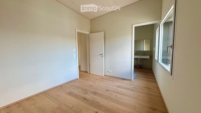 Empty room with light wooden flooring, white walls, and a doorway leading to a small bathroom with a sink and mirror.