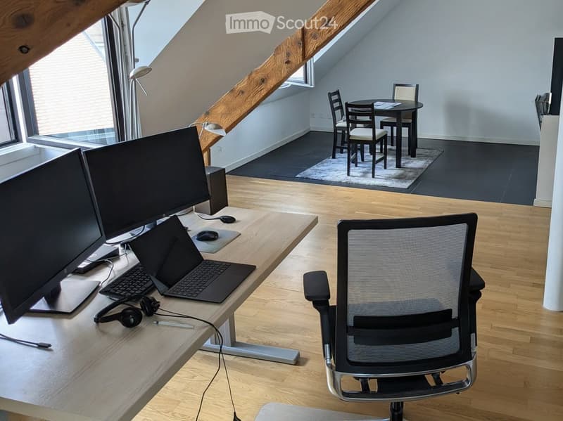 A modern, well-lit room with wooden flooring, a large desk with a laptop, dual monitors, and a comfortable office chair. A small dining area with a table and chairs is visible in the background.