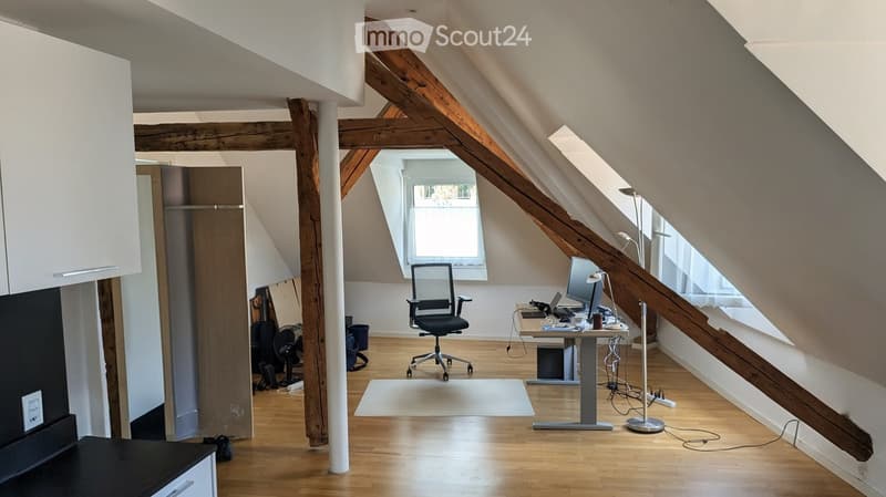 Spacious attic room with wooden beams, large windows, modern furniture including a desk, chair, laptop, and computer monitor.
