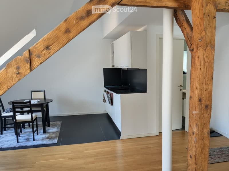 A modern, open-plan living and dining area with a black dining table and chairs, a sleek kitchen counter with cabinets, and exposed wooden beams on the ceiling.