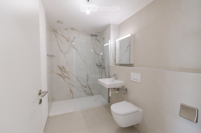 Modern bathroom with a white toilet, sink, and shower area featuring marble-patterned tiles. Includes a mirror with built-in lighting and a sleek glass shower enclosure.