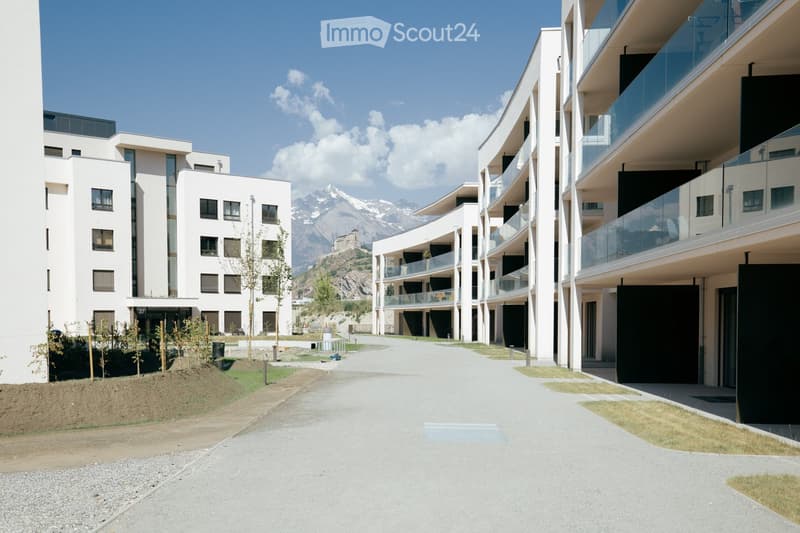 Modern residential complex with white buildings, balconies, and a scenic mountain backdrop.