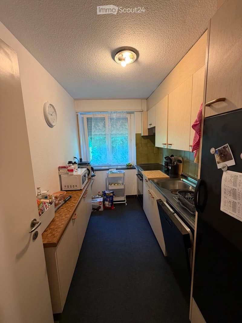 A compact kitchen with white cabinets, a dark refrigerator, microwave, oven, sink, and countertop. A small window provides natural light, and a clock is mounted on the wall.