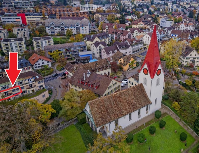 Church with red roof, surrounded by houses, trees, and grass, located in a city