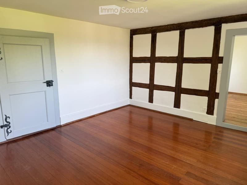 Empty room with wooden flooring, white walls, and a white door. Exposed wooden beams on one wall.
