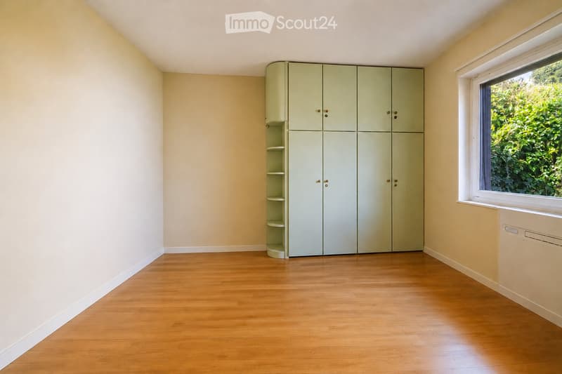 Empty room with light-colored walls, wooden flooring, and a large window with a view of greenery. A tall, light green wardrobe with shelves is positioned against one wall.