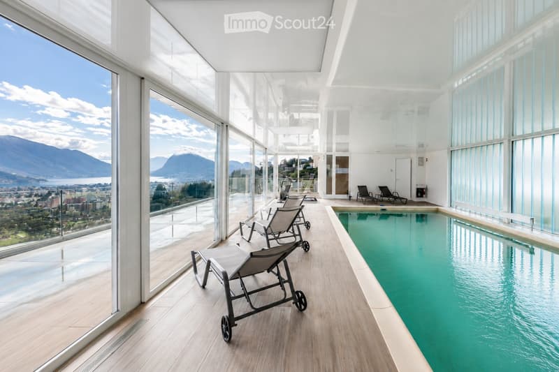 Indoor pool area with large windows offering a scenic mountain and lake view, featuring lounge chairs and a swimming pool.