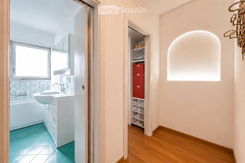 Modern bathroom with a white sink, bathtub, and a mirrored cabinet. The floor is tiled in turquoise, and the walls are painted in a light beige color. A wooden storage unit with red and white bins is visible in the adjacent hallway.