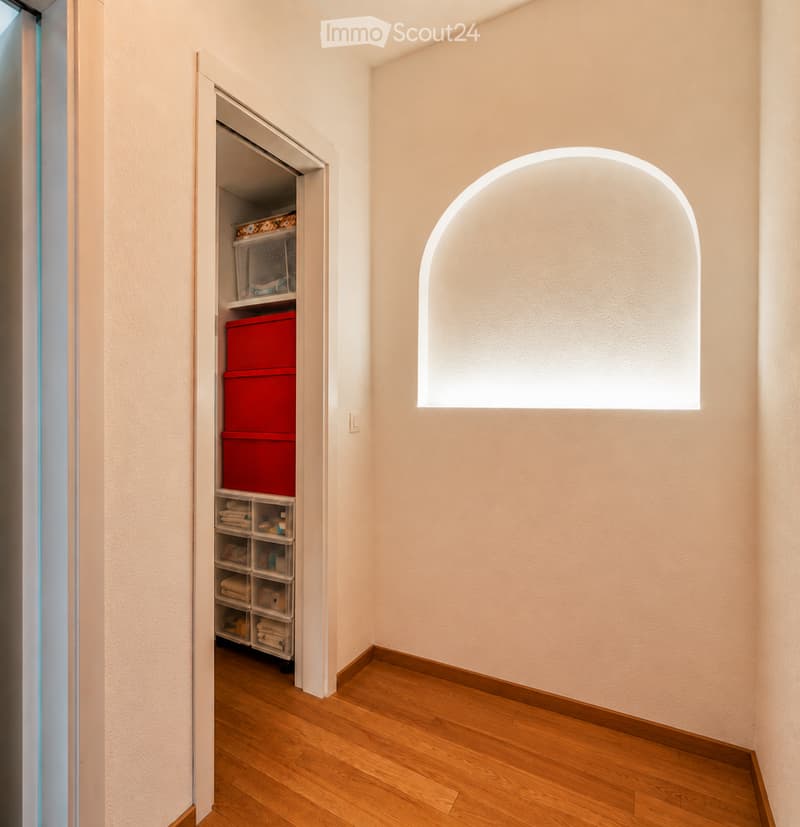 A narrow hallway with light-colored walls, wooden flooring, and an open closet with storage bins and folded towels.