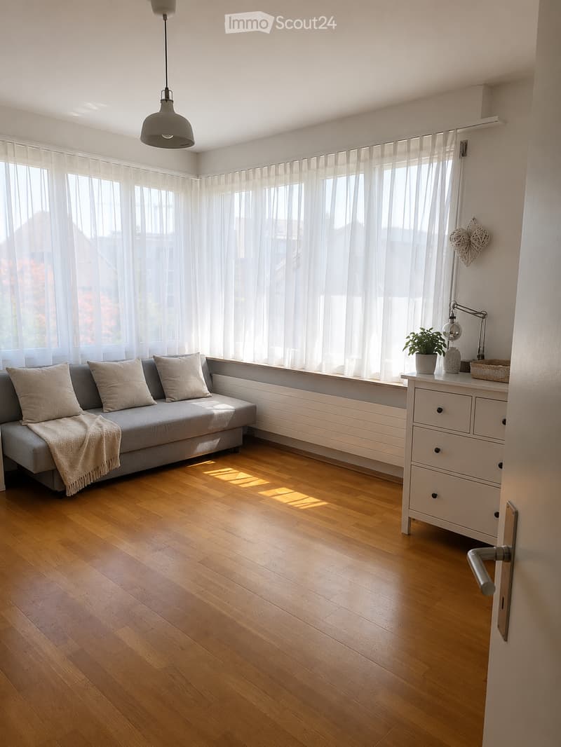 Modern living room with light wooden flooring, grey sofa with throw blanket, side table with lamp and plant, large windows with sheer curtains, and a hanging pendant light.