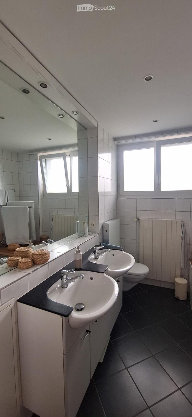 Modern bathroom with two sinks, a toilet, a large mirror, and a towel rack. The countertop features a black surface and a soap dispenser, with wicker baskets on the left side.