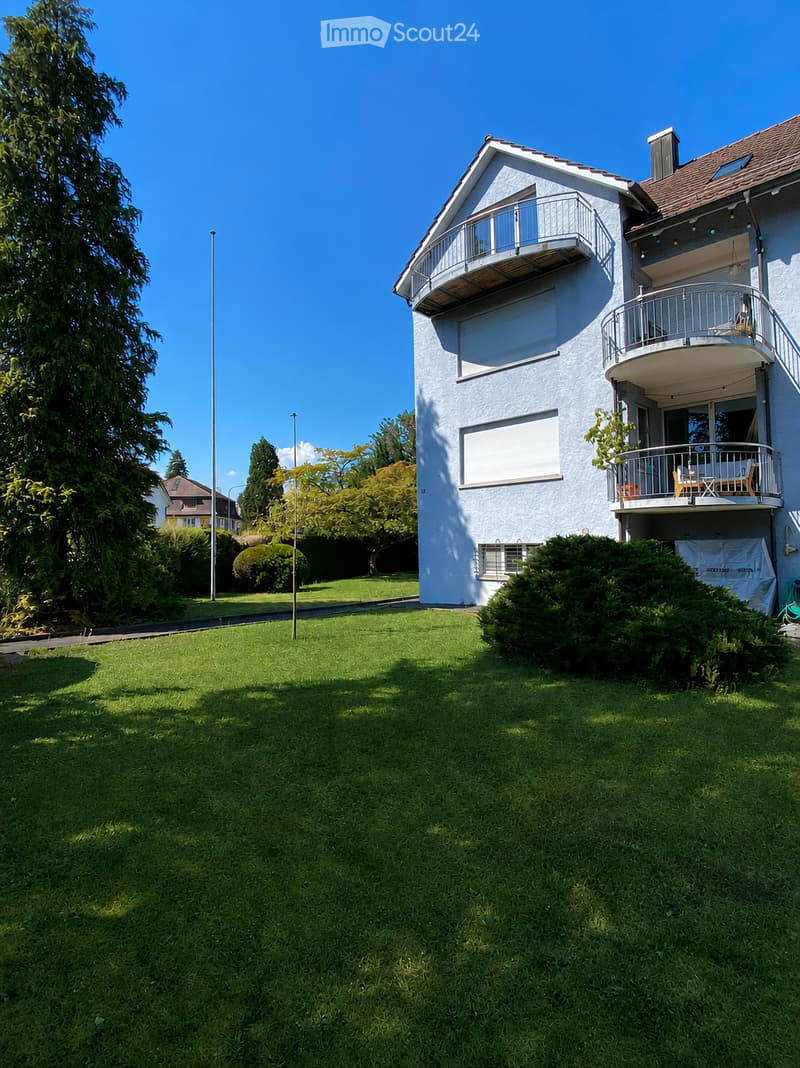 A modern two-story house with white exterior and multiple balconies, surrounded by a well-maintained lawn and trees.