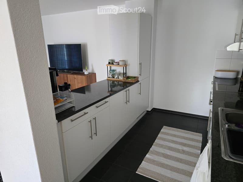 Modern kitchen with white cabinets, black countertops, and stainless steel fixtures. Includes a sink, wooden shelf, and small table with a screen.