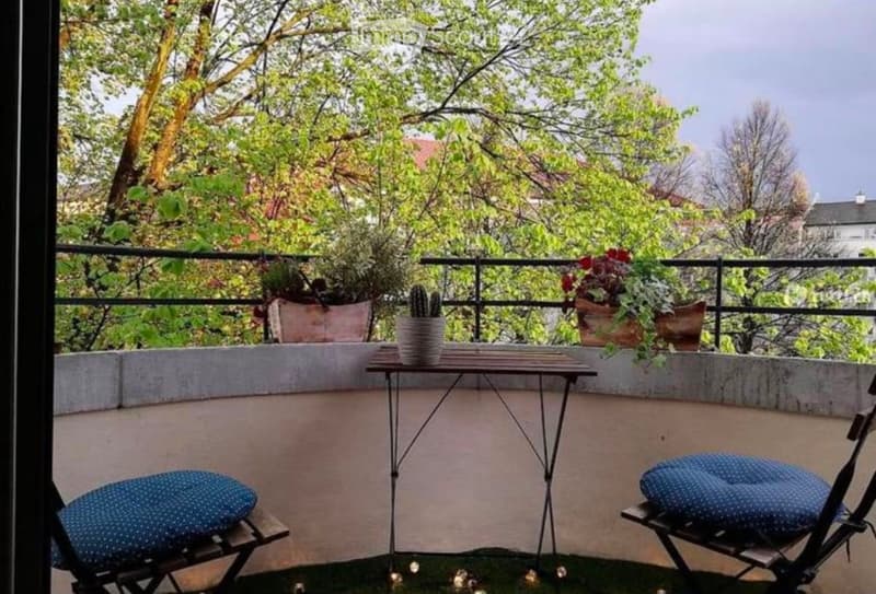 Balcony with a small table, two chairs, and potted plants, surrounded by greenery and trees.