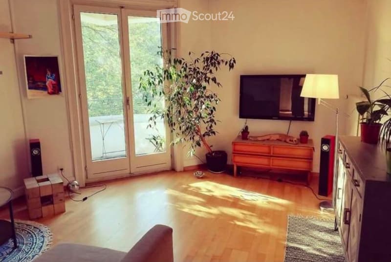 Bright living room with wooden flooring, large sliding glass doors leading to a balcony, a sofa, a coffee table, a TV mounted on the wall, a floor lamp, and several plants.