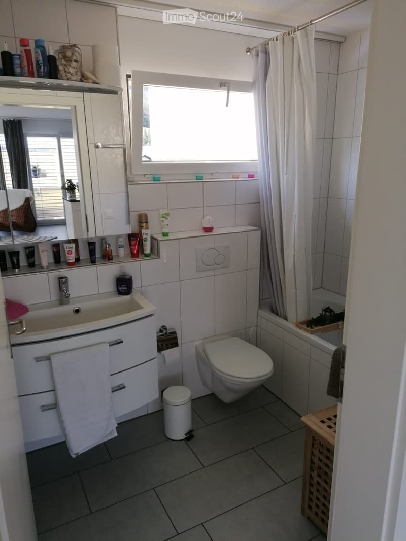Modern bathroom with white tiles, a sink, mirror, toilet, bathtub, and various toiletries on the counter. Features a window with blinds and a small wooden shelf near the bathtub.