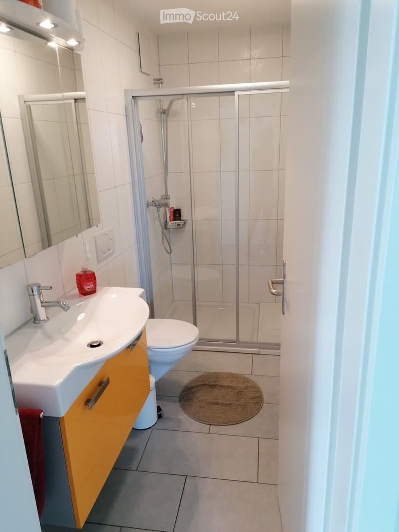 Modern bathroom with white tiles, a white sink with a silver faucet, a toilet, a glass-enclosed shower, a mirror, and a round bath mat on the floor.