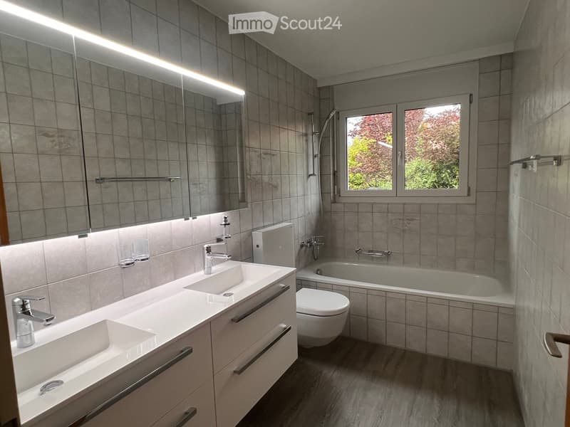 Modern bathroom with double sink, toilet, bathtub, large mirror, and window showing greenery outside.