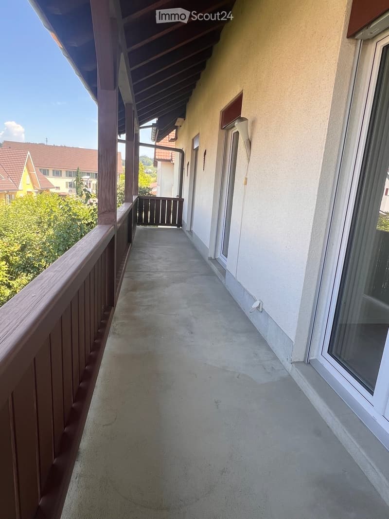 Balcony with wooden railing, concrete floor, and a view of greenery and neighboring houses.