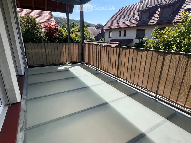 Balcony with a modern glass floor and wooden privacy screens, overlooking a residential area with houses and trees.