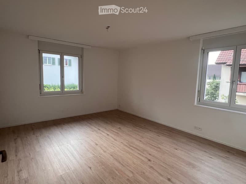 Empty room with light wooden flooring and white walls, featuring two windows with white frames and blinds.
