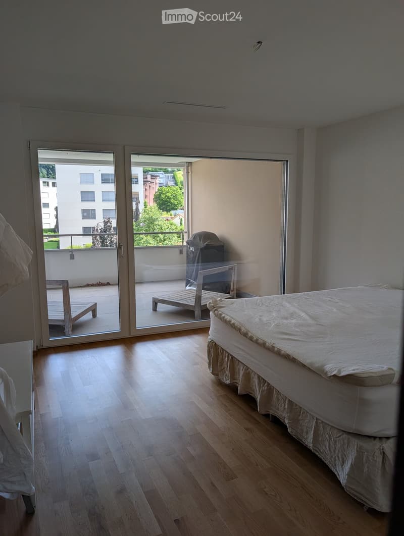 Bright bedroom with wooden flooring, a neatly made bed, and a sliding glass door leading to a balcony with outdoor furniture.