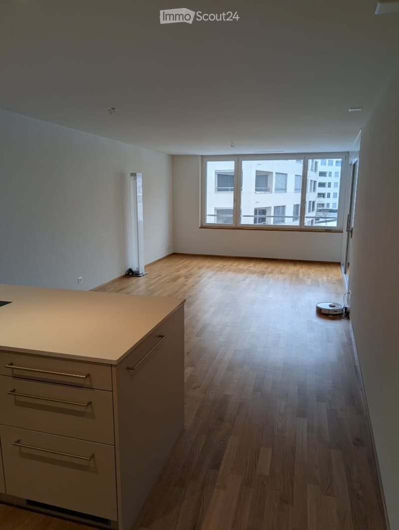 Open-plan space with a modern kitchen island and a tall floor lamp near the window. The room features wooden flooring and large windows offering a view of the city.