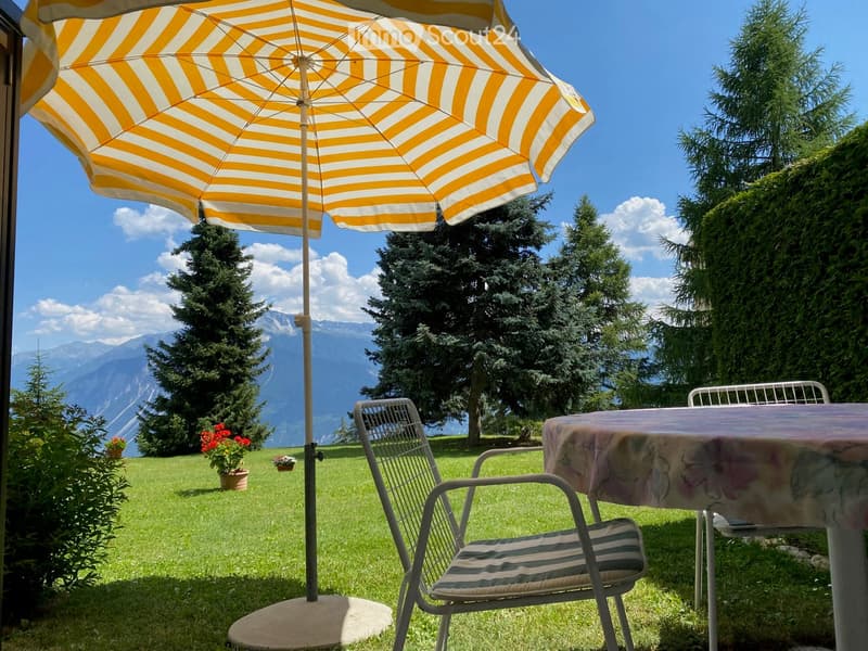 Garden with a patio umbrella, table, and chairs, surrounded by greenery and a mountain view.