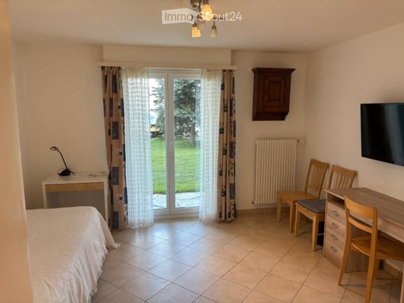 Cozy furnished room with a bed, desk, chair, table, radiator, cabinet, lamp, curtain, and screen. The room has a view of a green outdoor area through large glass doors.