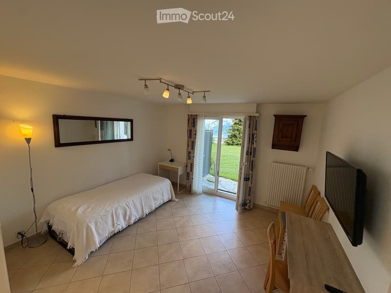 A furnished room with a bed, a small table, chairs, a mirror, a floor lamp, and a television. The room has tiled flooring and a sliding glass door leading to a grassy outdoor area.