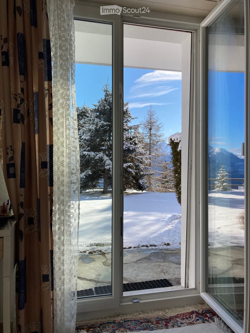 Large window with a view of snow-covered trees and mountains, partially covered by curtains and a lace panel.