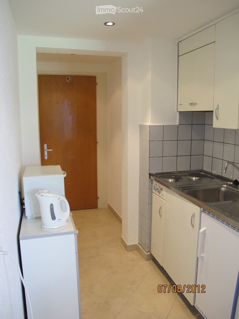 Modern kitchen with white cabinets, stainless steel sink, electric kettle, microwave, and refrigerator. The floor is tiled, and the walls are white.