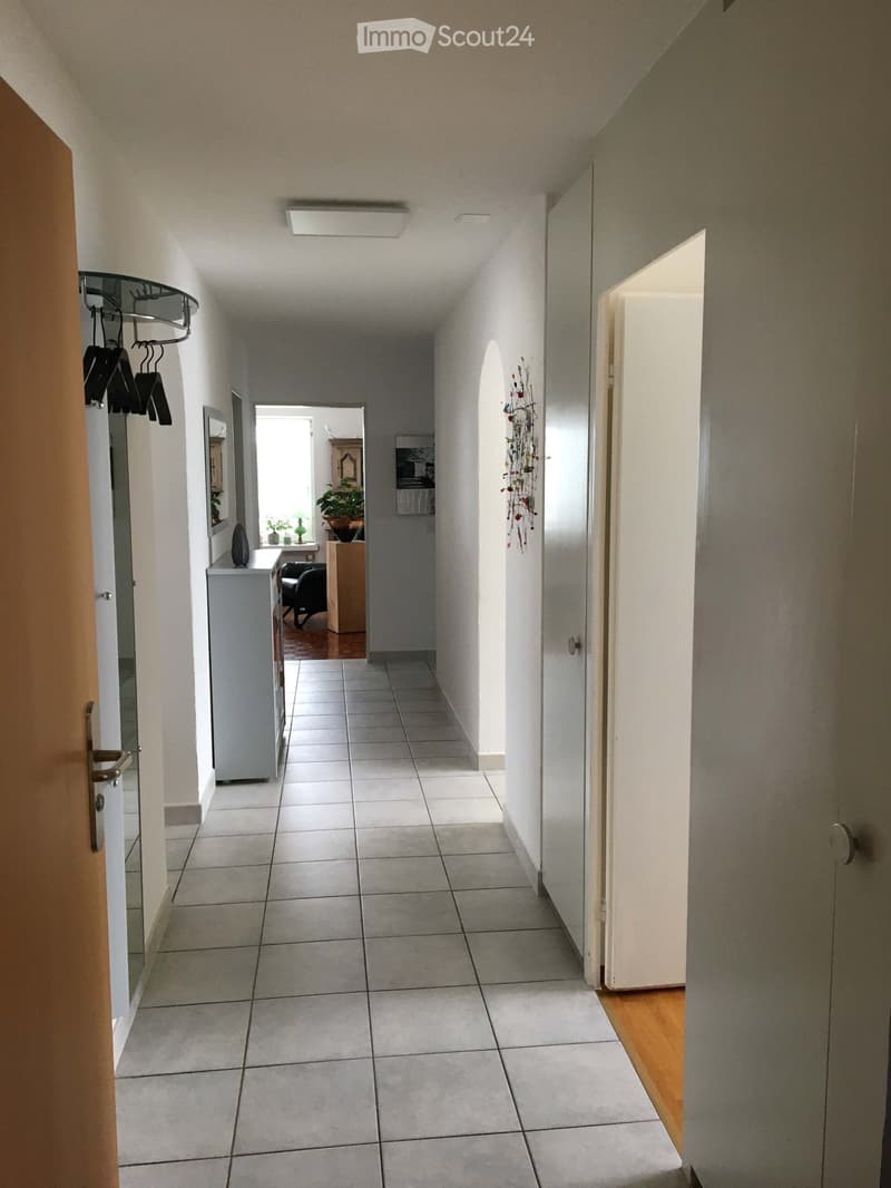 A clean, tiled hallway with white walls, a coat rack, and an open door leading to another room.