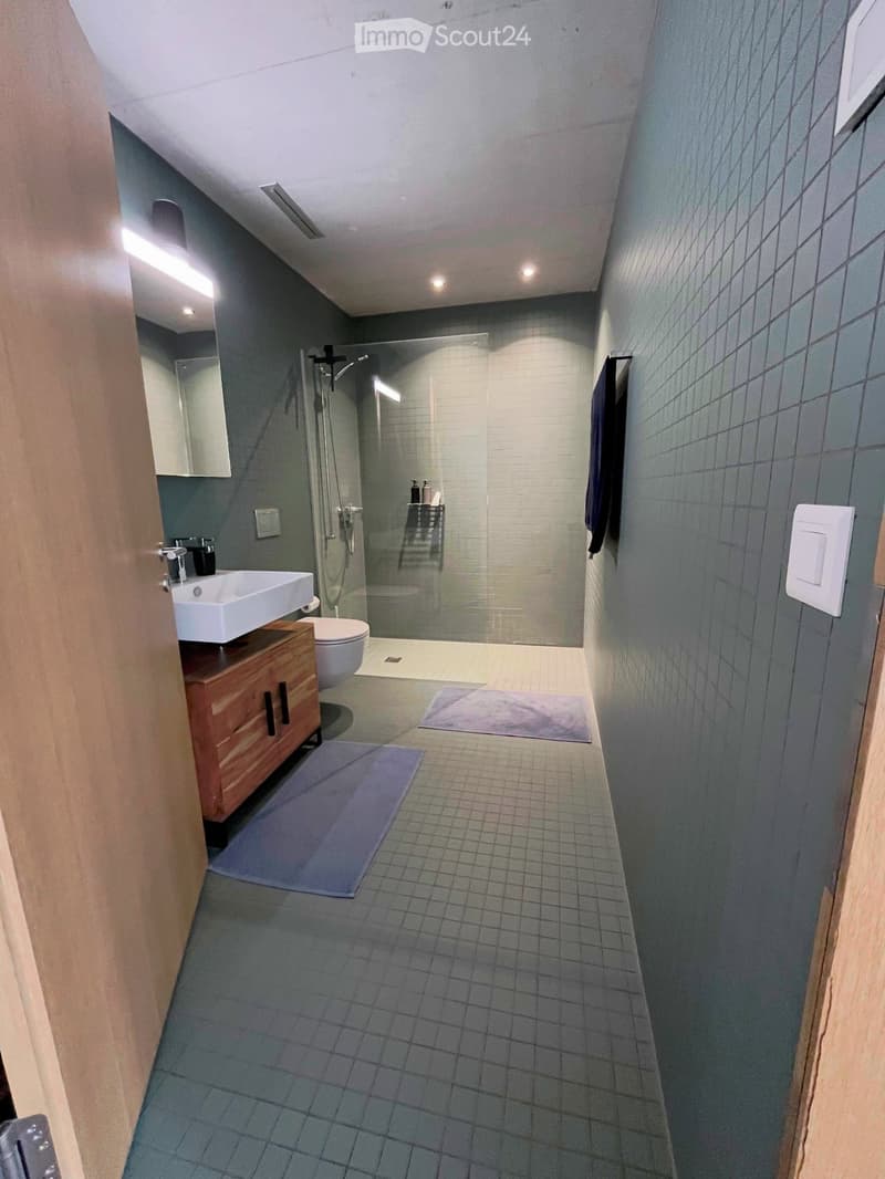 Modern bathroom with grey tiled walls and floor, white sink, toilet, shower, and wooden cabinet.