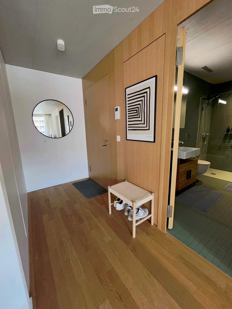 Hallway with wooden flooring, a small woven bench holding shoes, a round mirror, and an open door leading to a modern bathroom with a sink, toilet, and glass shower enclosure.