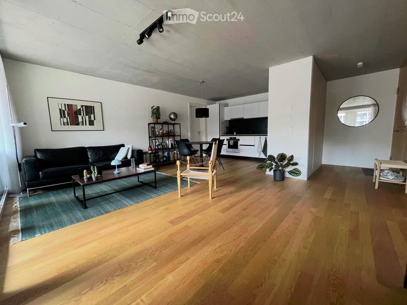 Open-plan living and dining area with wooden flooring, a black leather sofa, a coffee table, a dining table with chairs, and a modern kitchen in the background.