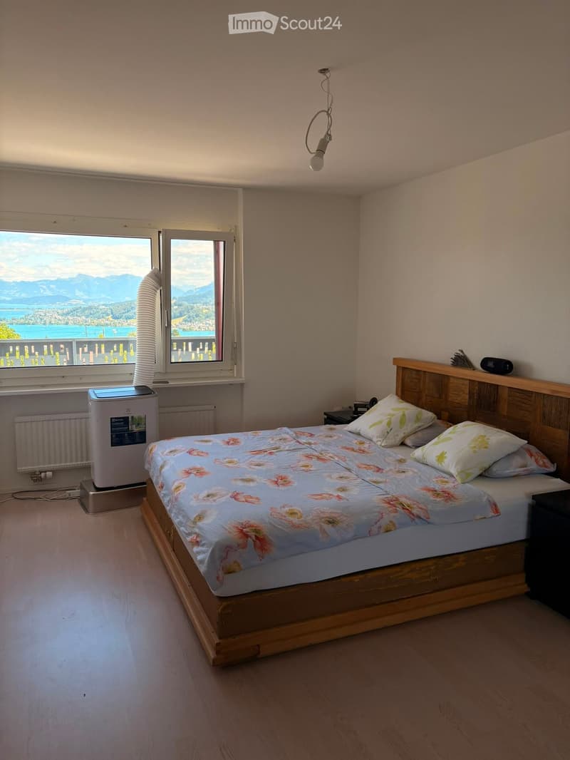 Cozy bedroom with wooden bed, floral bedding, nightstand, radiator, and air conditioner. Large window offers scenic mountain and lake view.