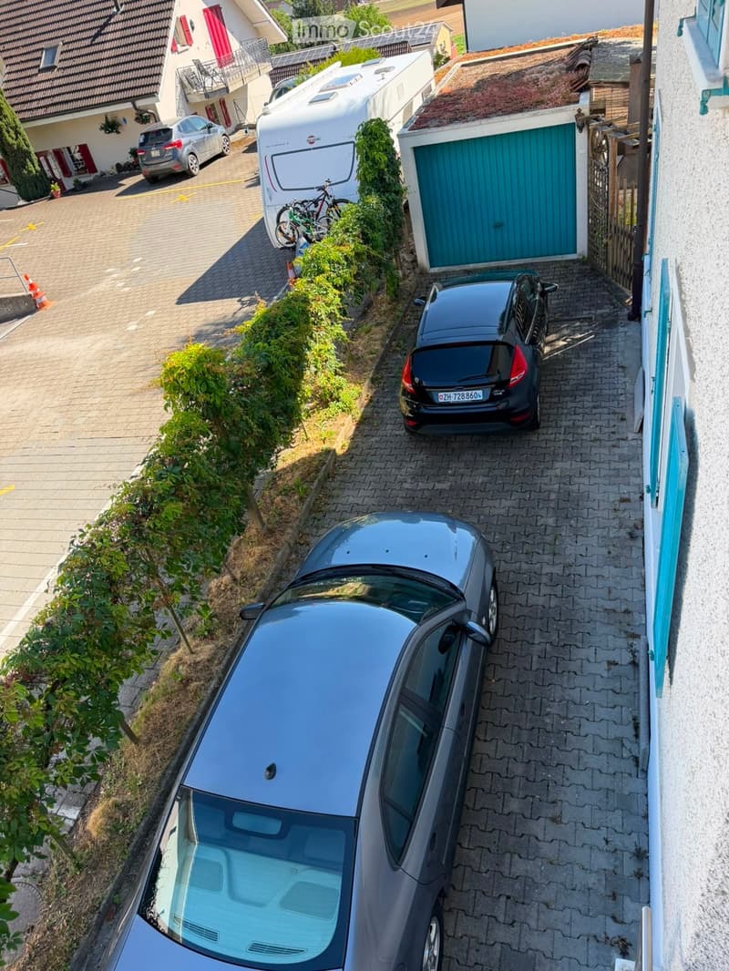 Aerial view of a residential area showing a driveway with parked cars, a garage, and neighboring houses with balconies and solar panels.